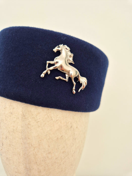 Navy blue felt hat with a silver horse brooch on a mannequin head against a beige background close up