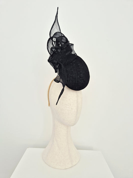 A black straw percher fascinator with a tall pointed knotted detail and a round structured base, displayed on a cream-colored mannequin head against a plain white background. side view