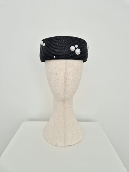 A black straw headpiece with white faux-pearl embellishments, displayed on a cream-colored mannequin head against a plain white background.