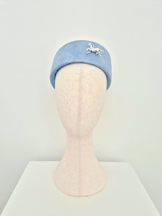 A dusty blue straw pillbox headpiece worn towards the back with a silver horse brooch on the front, displayed on a beige mannequin head against a white background.