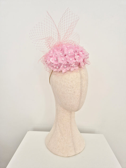 A light pink fascinator hat with a floral crown and sheer netting, displayed on a cream-colored felt mannequin head against a plain white background.side view