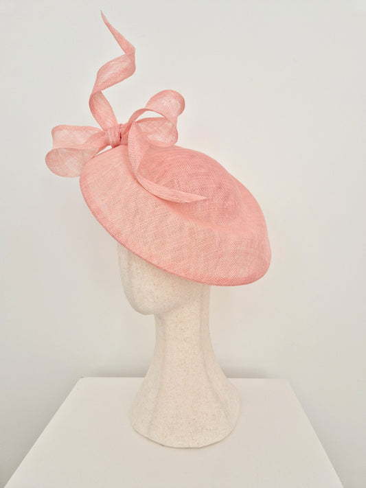 A light pink fascinator hat with a large matching ribbon bow, displayed on a textured off-white mannequin head and stand against a plain white background.side view