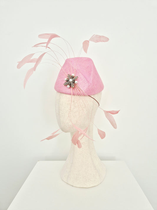 A pink fascinator hat with delicate feather accents and a jeweled center, displayed on a textured cream mannequin head against a clean white background. side view