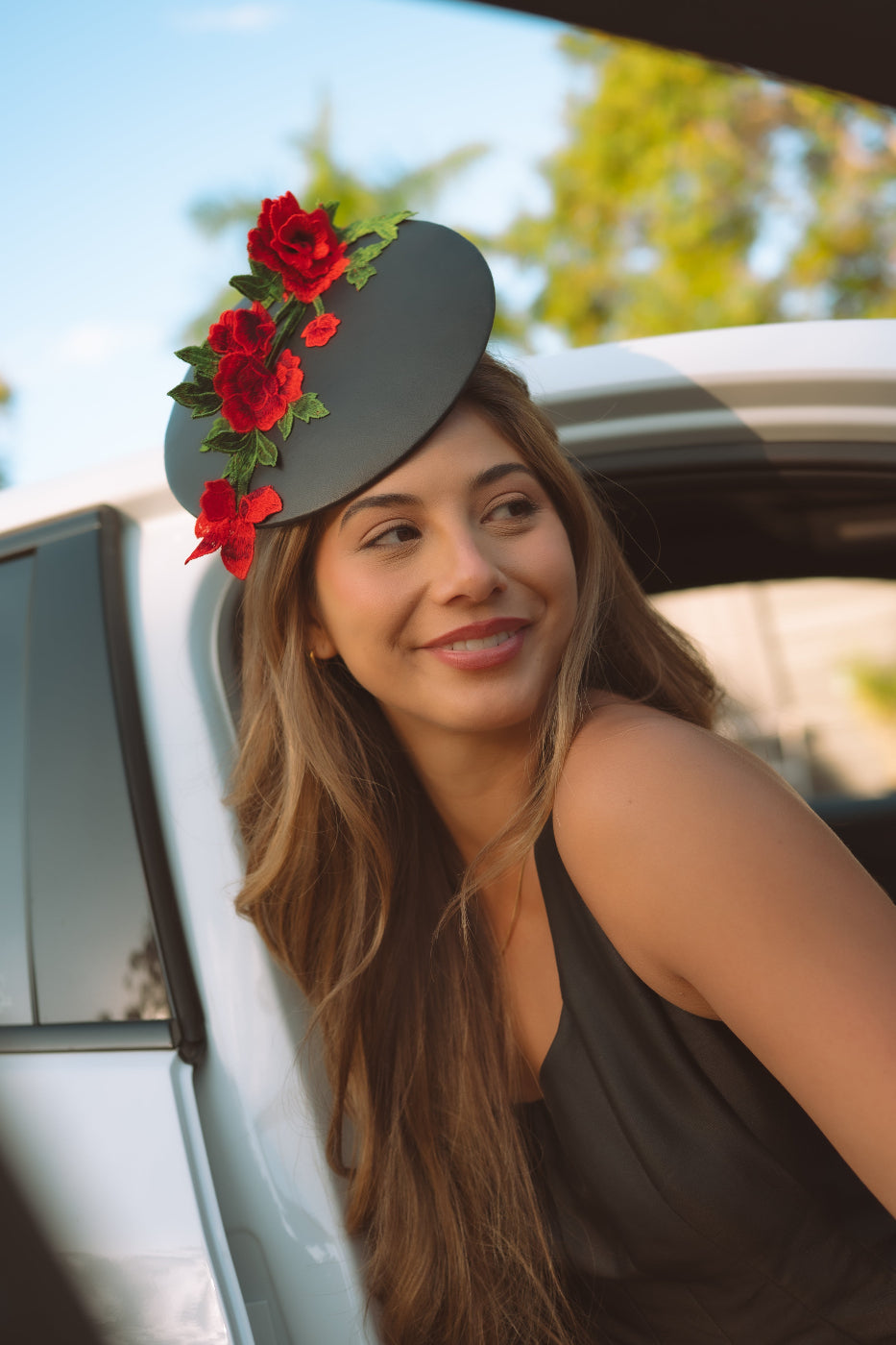 A woman leaning out of a white car wearing a black leather fascinator hat adorned with red roses and green leaves.