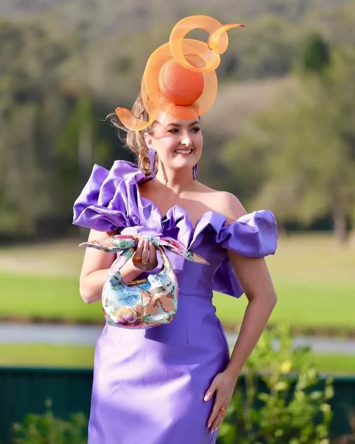 Woman in a vibrant purple dress with a colorful hat, holding a floral bag, outdoors.