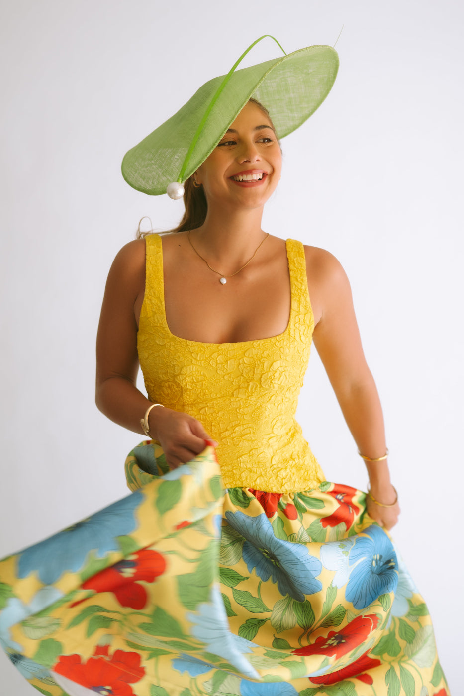 A woman wearing a bright yellow sleeveless lace bodice top and a voluminous yellow skirt with a colorful floral print, paired with a wide-brimmed light green fascinator hat, posing against a white studio background.
