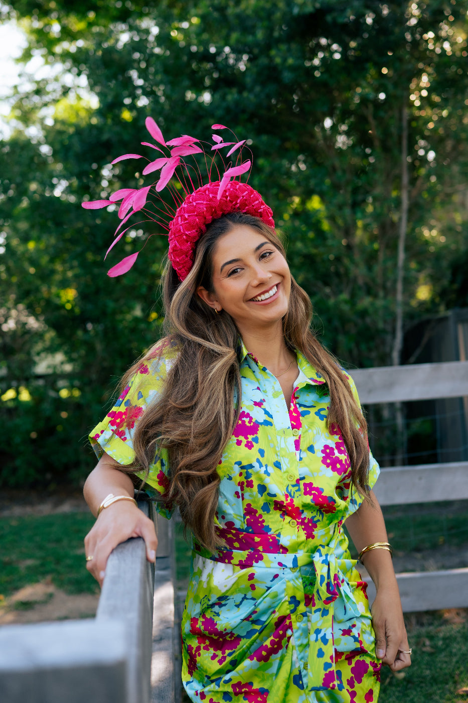 A woman outdoors wearing a bright lime green floral dress with pink, blue, and yellow patterns, paired with a matching pink fascinator headpiece. She is smiling and leaning on a wooden fence with a lush green garden background.