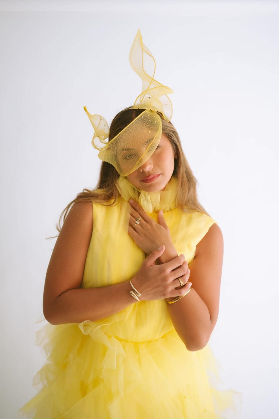 A woman wearing a bright yellow sleeveless dress with a ruffled tulle skirt and a matching yellow fascinator headpiece, posed against a plain white studio background.