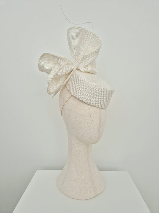 An off-white textured straw fascinator hat with a large bow, displayed on a matching off-white felt mannequin head against a plain light gray background. side view