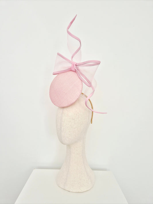 A light pink straw percher fascinator headpiece with a large bow on a mannequin head against a white background. side view