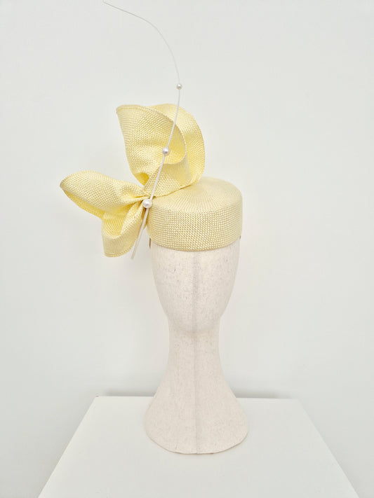 A pale yellow straw pillbox fascinator hat with a scrunch bow and white quill detail, displayed on a textured off-white mannequin head against a clean white background.