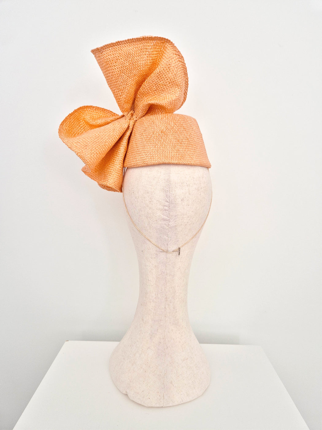 An peach straw fascinator hat with a large bow, displayed on a light beige mannequin head against a white background. back view