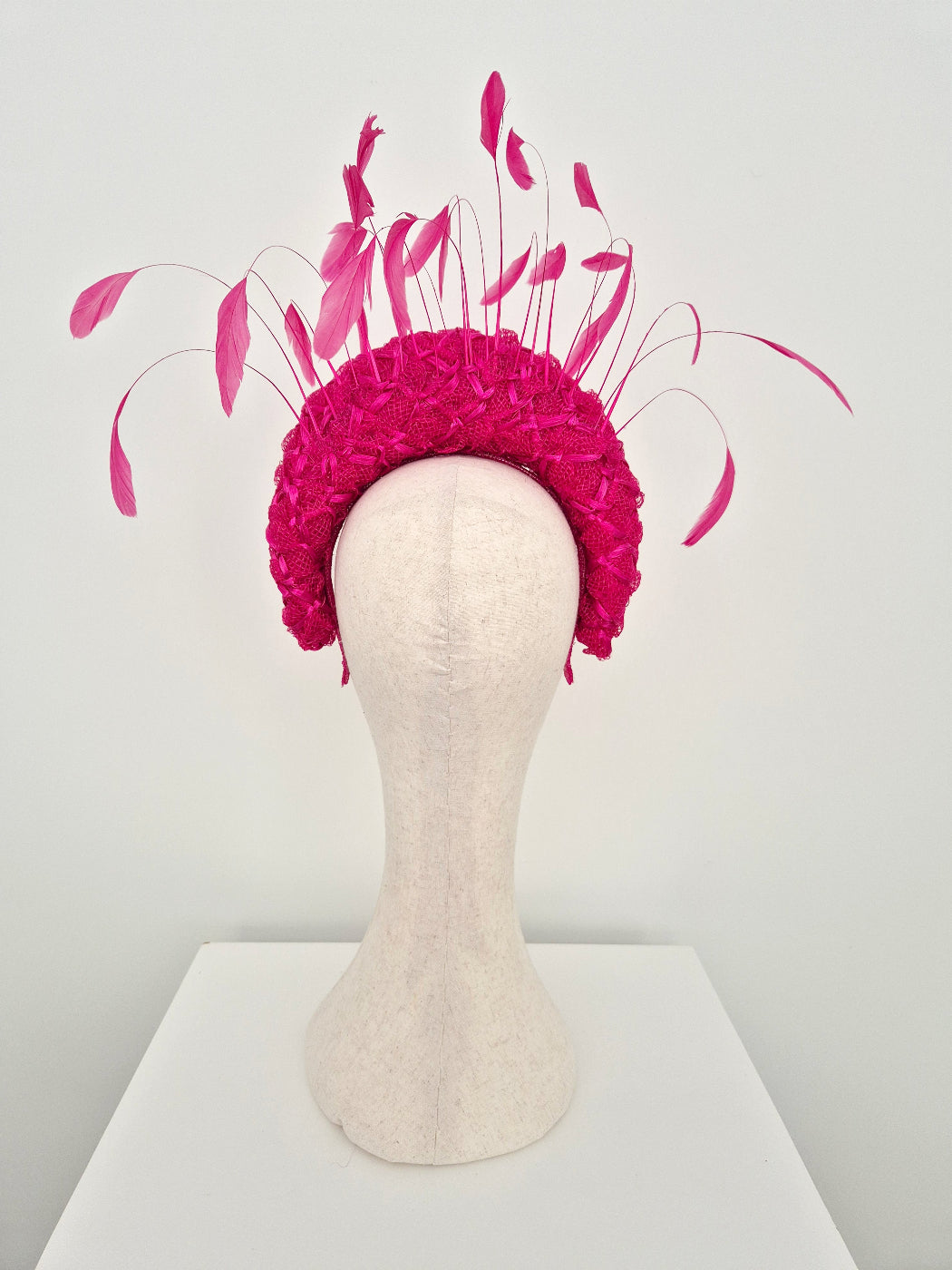 A raspberry pink fascinator headpiece with long feather accents, displayed on a cream fabric mannequin head against a plain light background. back view