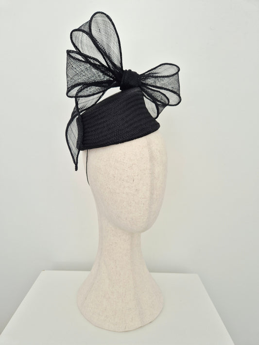 A black straw fascinator with a large black mesh bow, displayed on a textured off-white mannequin head against a clean white background side front view
