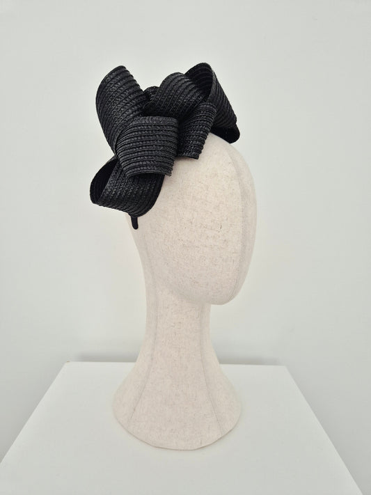 A black braided loop fascinator hat , displayed on a textured off-white mannequin head against a clean white background. side view