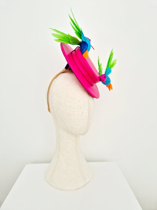 A bright pink fascinator hat displayed on a textured off-white mannequin head. The hat features a wide pink brim and is decorated with two colorful bird embellishments in blue, green, and orange, set against a plain white background. side view