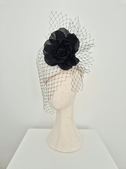 A white straw fascinator with a large fabric rose and matching black birdnet veil, displayed on a cream-colored felt mannequin head against a plain white background.side view