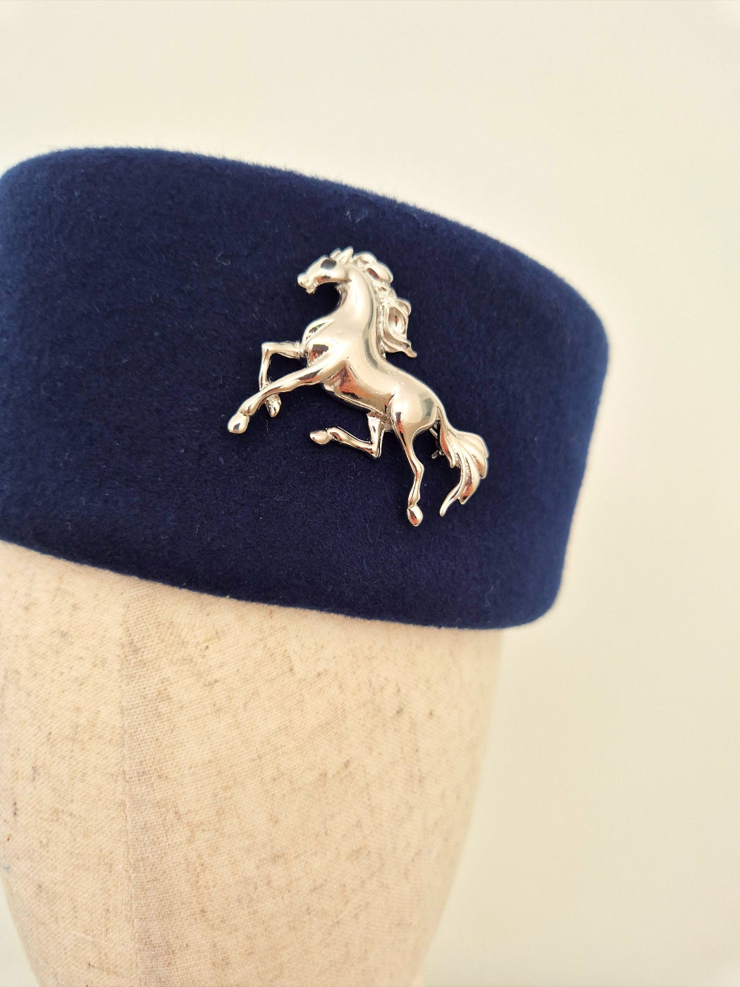 Navy blue felt hat with a silver horse brooch on a mannequin head against a beige background close up