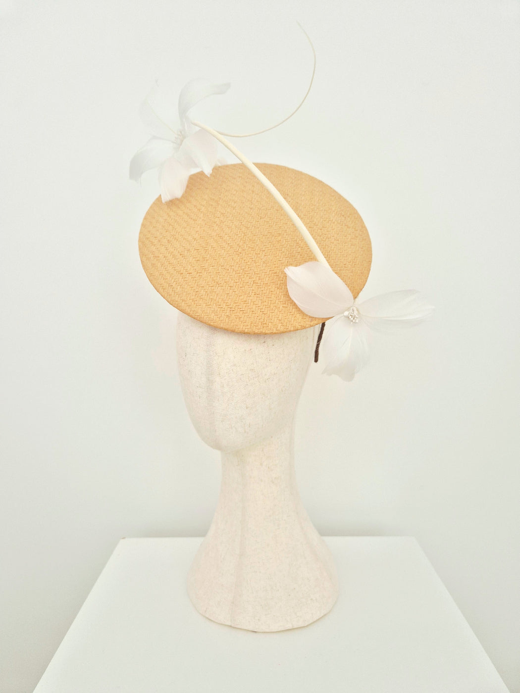 A light beige woven straw percher fascinator headpiece with white feather flowers and a curved feather accent, displayed on a textured cream mannequin head against a white background. side view