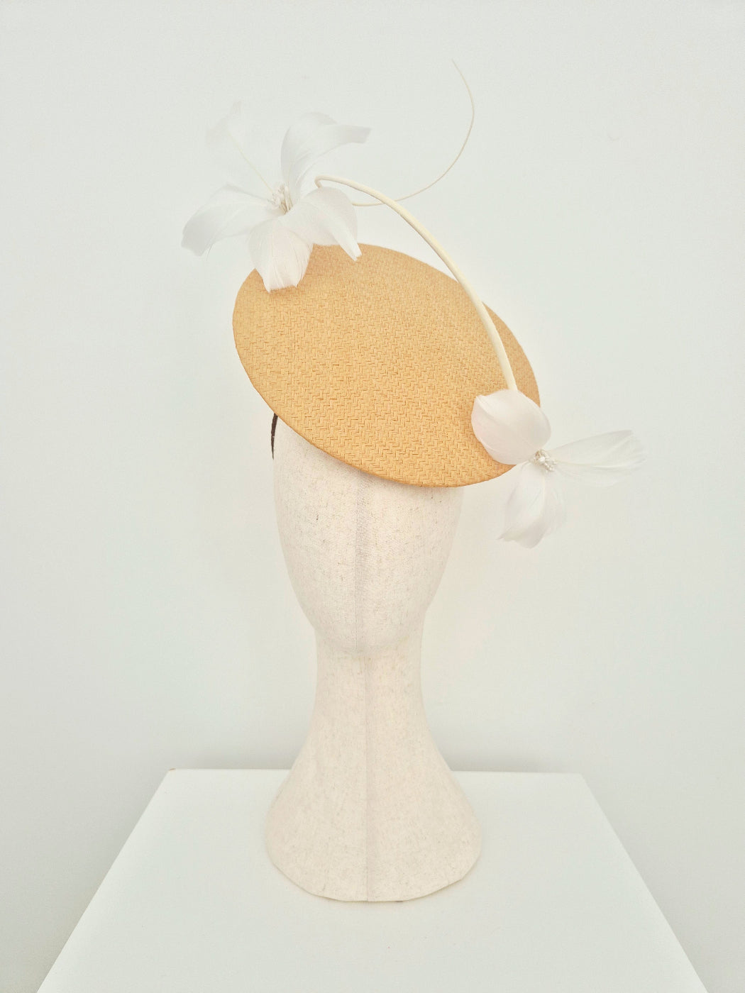 A light beige percher fascinator with white feather flowers and a long feather accent, displayed on a textured cream mannequin head against a white background.