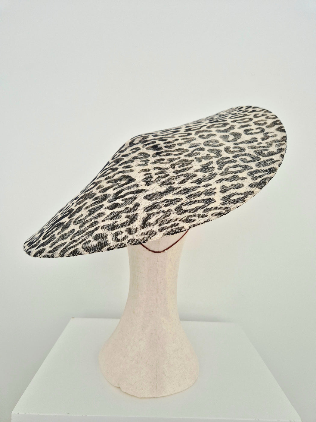A wide-brimmed  hat black and beige leopard print pattern, displayed on a textured off-white mannequin stand against a plain white background. back view