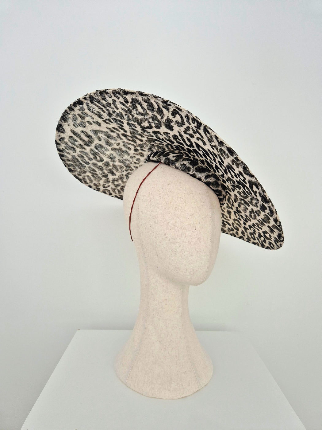 A wide-brimmed fascinator hat with a black and beige leopard print pattern, displayed on a cream-colored mannequin head against a white studio background.underside 2
