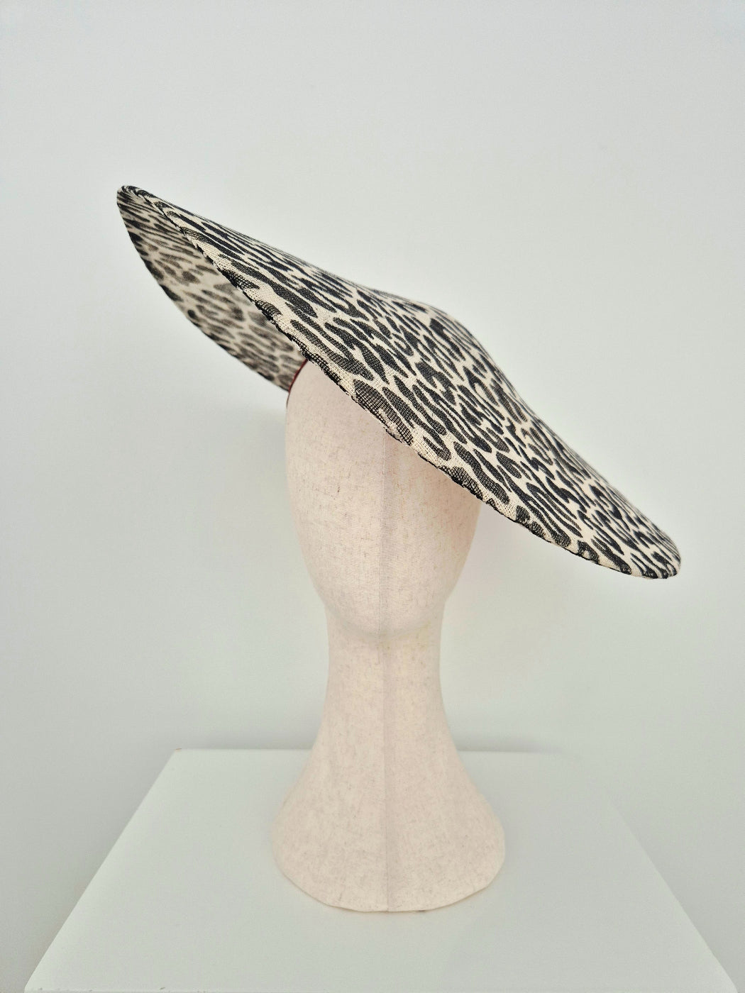A wide-brimmed fascinator hat with a black and beige leopard print pattern, displayed on a light pink mannequin head against a white studio background.