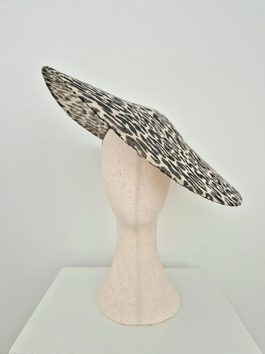 A wide-brimmed fascinator hat with a black and beige leopard print pattern, displayed on a light pink mannequin head against a white studio background.