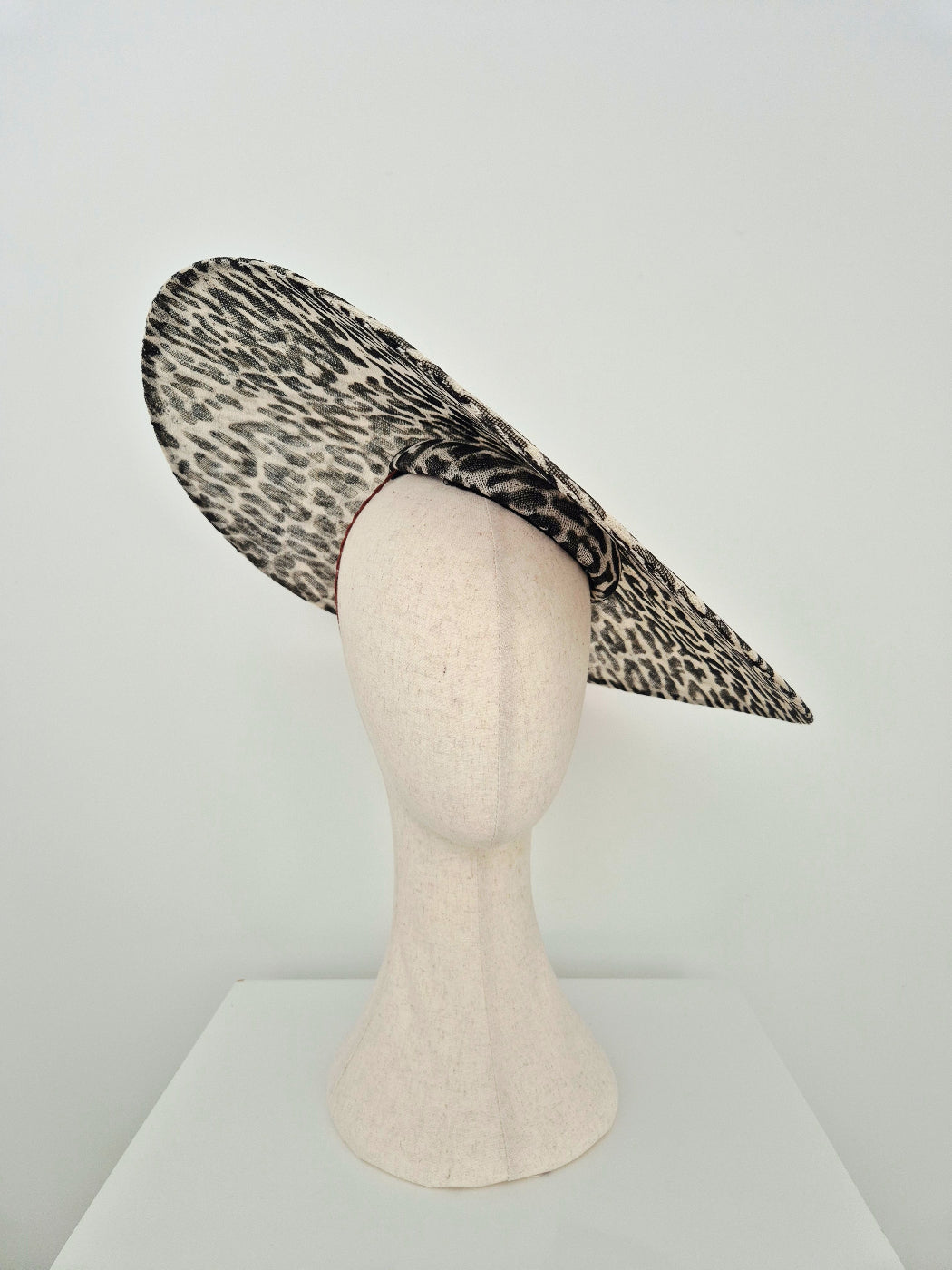 A wide-brimmed fascinator hat with a black and beige leopard print pattern, displayed on a textured off-white mannequin head against a plain white background.underside