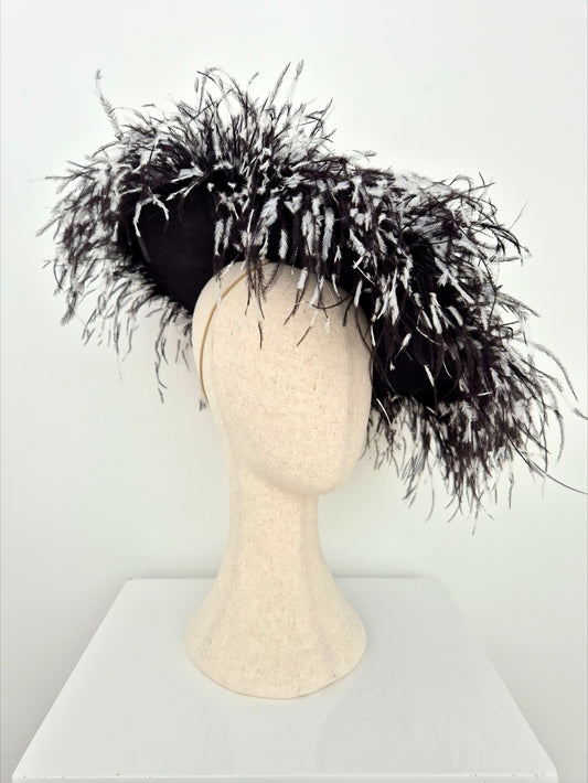 A black fascinator hat with dramatic black-and-white feathered trim, displayed on a cream mannequin head against a plain white background.side view
