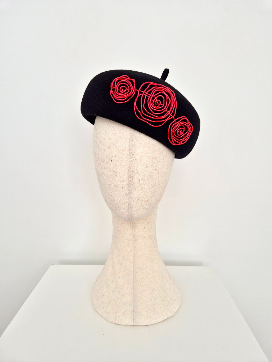 A black felt beret with three red wire roses, on a mannequin head against a white background.