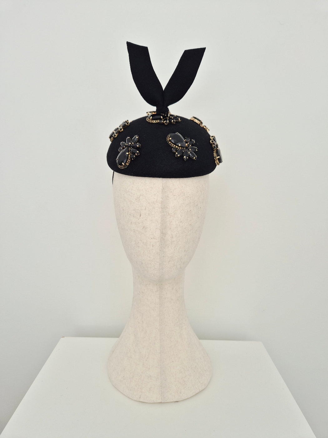A black felt hat with rhinestone embellishments and a black ribbon bow, displayed on a cream-colored mannequin head against a white background.front on