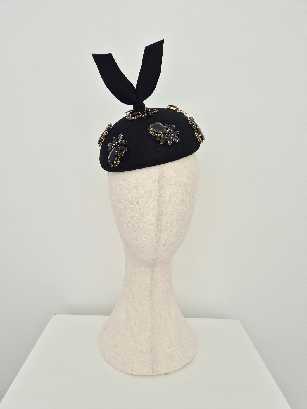 A black felt headpiece with rhinestone embellishments and a black ribbon bow, displayed on a cream-colored mannequin head against a light gray background.