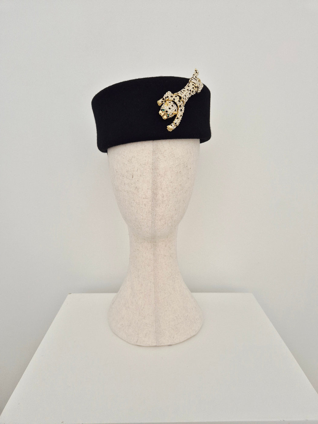 Black felt pillbox with a gold and rhinestone leopard embellishment on a mannequin head against a white background