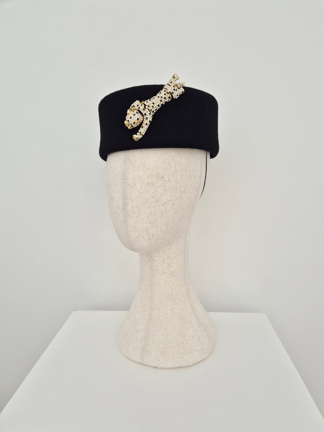 Black pillbox headpiece with decorative brooch on a mannequin head against a white background side view
