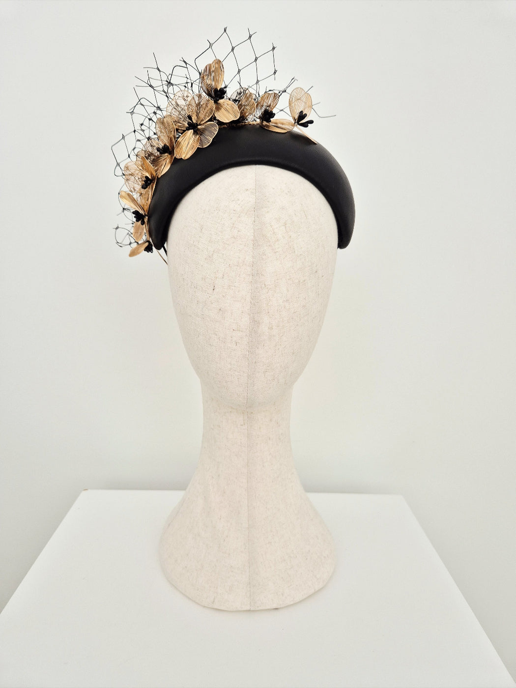 Black bandeau headpiece with gold floral decorative elements on a mannequin head against a white background
