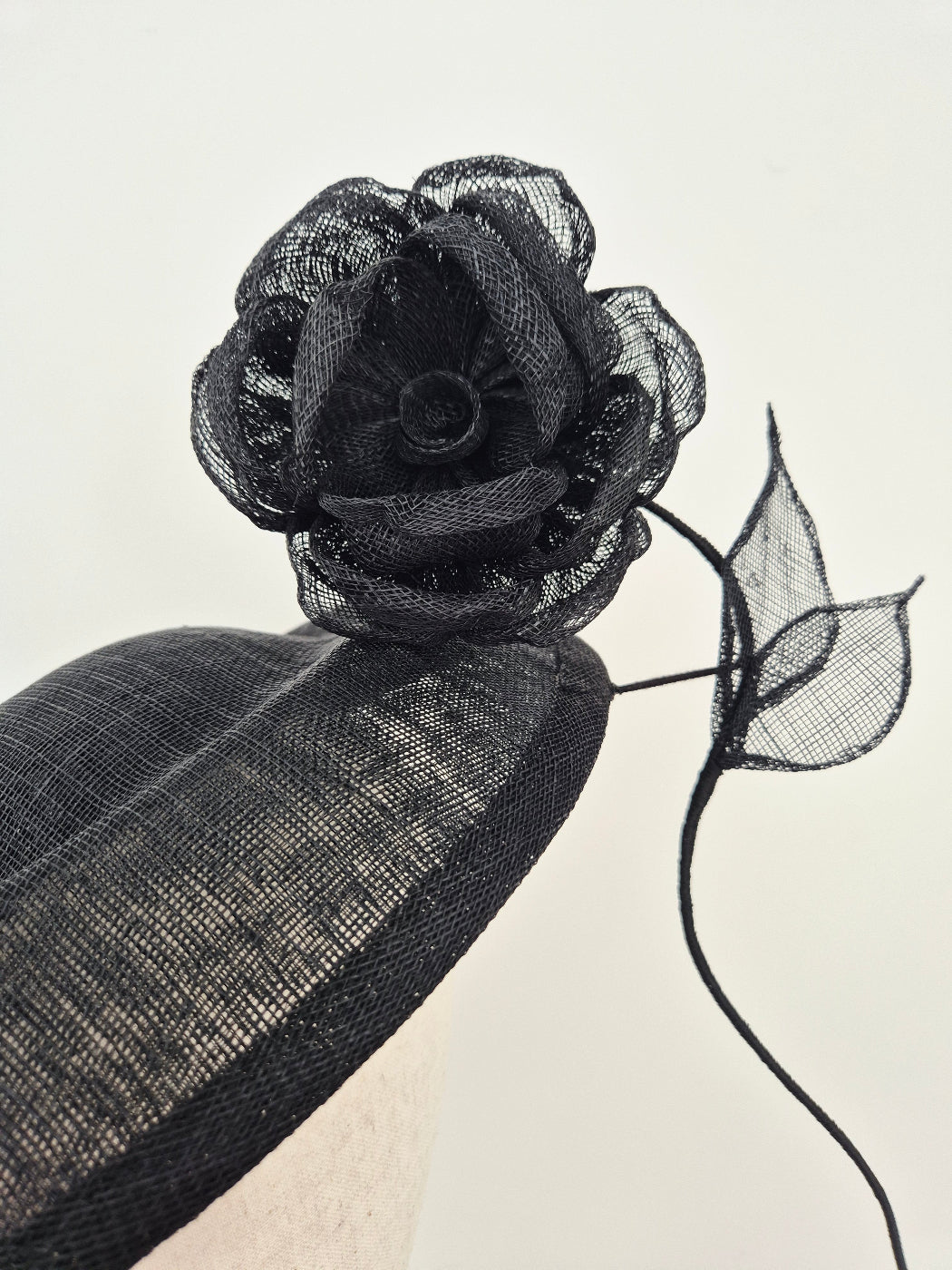 A close-up of a black fascinator hat featuring a sheer mesh flower with leaf-like accents and a structured black fabric base, shown against a plain light background.