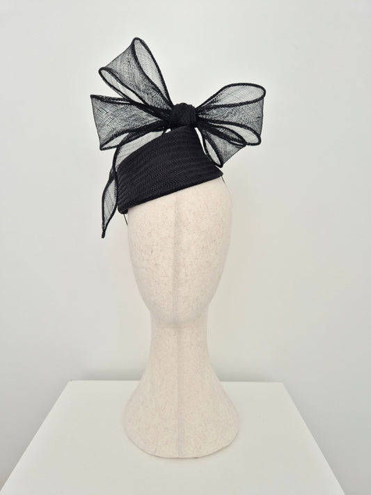 A black fascinator with a large bow displayed on a cream-colored mannequin head against a minimalist white background.