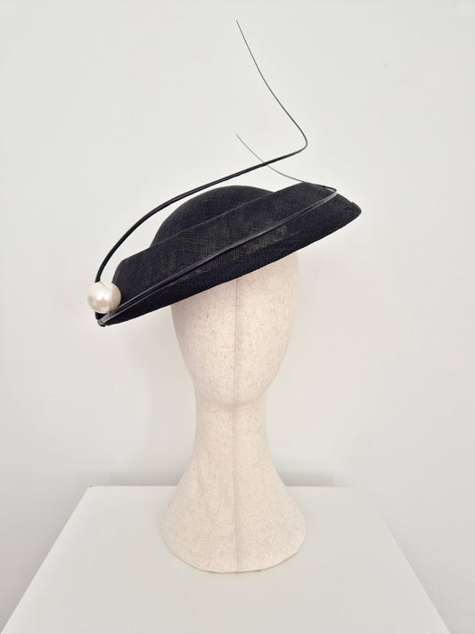 A black straw fascinator hat with a curved black quills and a single pearl bead, displayed on a cream-colored felt mannequin head against a white background.