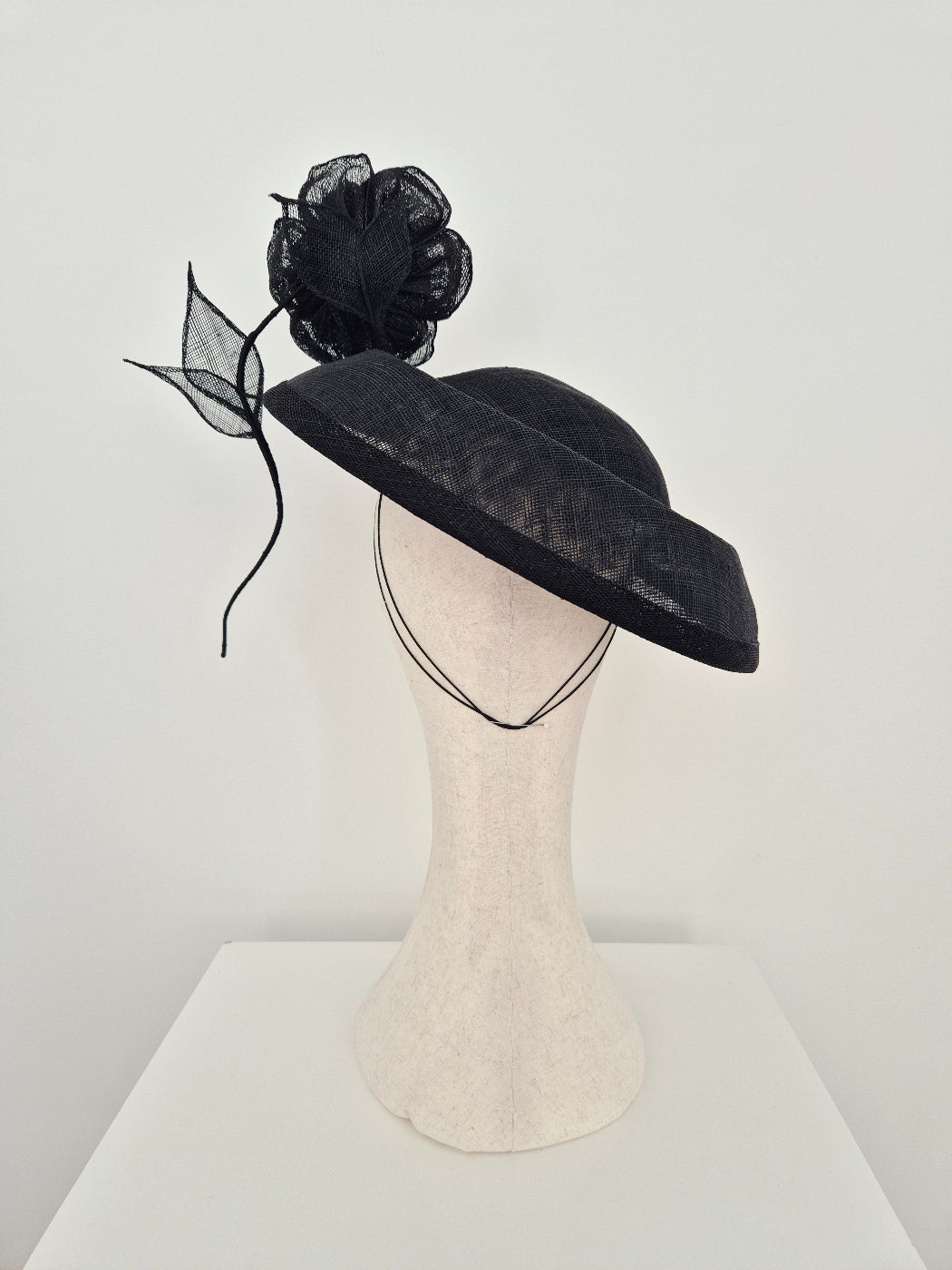 A black fascinator hat with a sheer black flower and leaf accent, displayed on a cream-colored mannequin head against a minimalist white background.back view