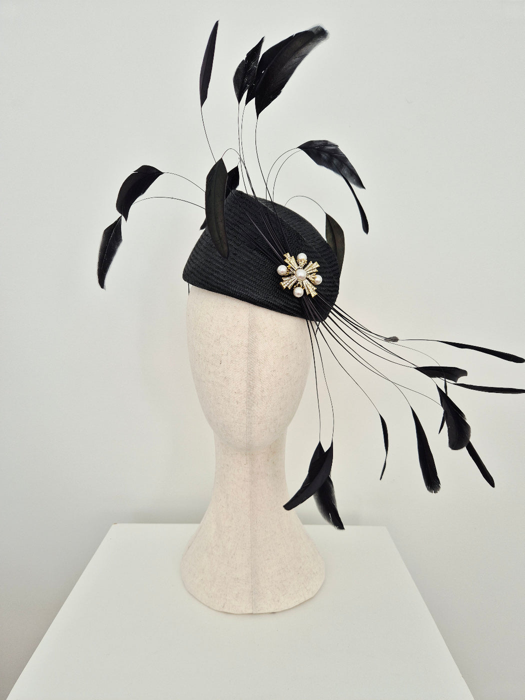 A black fascinator hat with long feather plumes and a pearl-like floral brooch, displayed on a cream-colored mannequin head against a light gray wall.