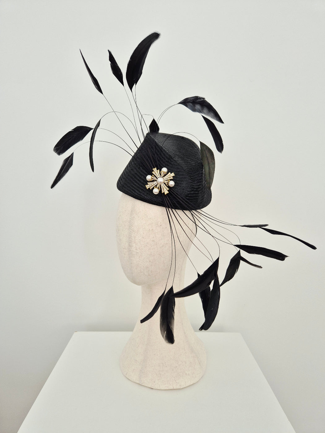 A black fascinator hat with a textured black crown, a white floral brooch, and long black feather plumes, displayed on a cream-colored mannequin head against a light gray wall. side view