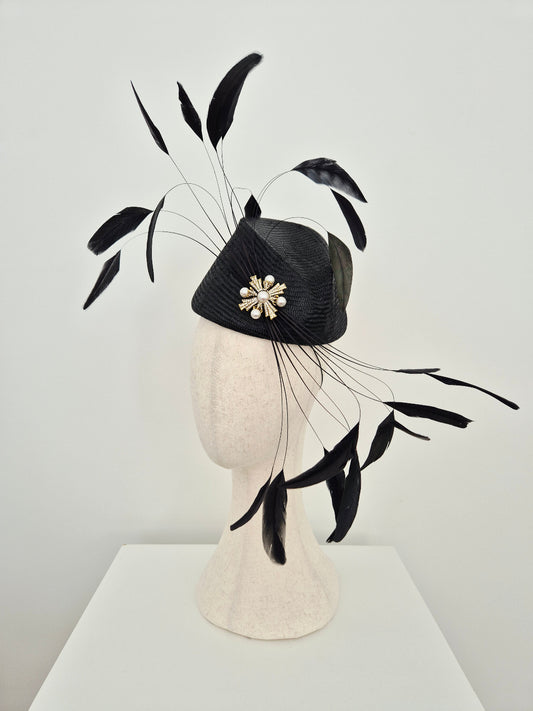 A black fascinator hat with a textured black crown, a white floral brooch, and long black feather plumes, displayed on a cream-colored mannequin head against a light gray wall. side view