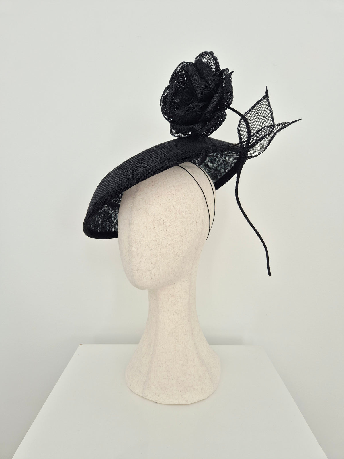 A black fascinator hat with a large black mesh rose and matching mesh leaf accent, displayed on a cream-colored mannequin head against a plain white background.side view