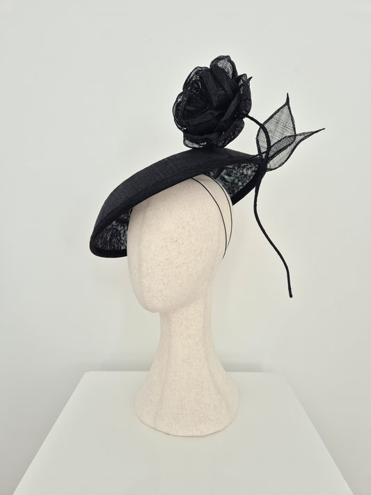 A black fascinator hat with a large black mesh rose and matching mesh leaf accent, displayed on a cream-colored mannequin head against a plain white background.side view