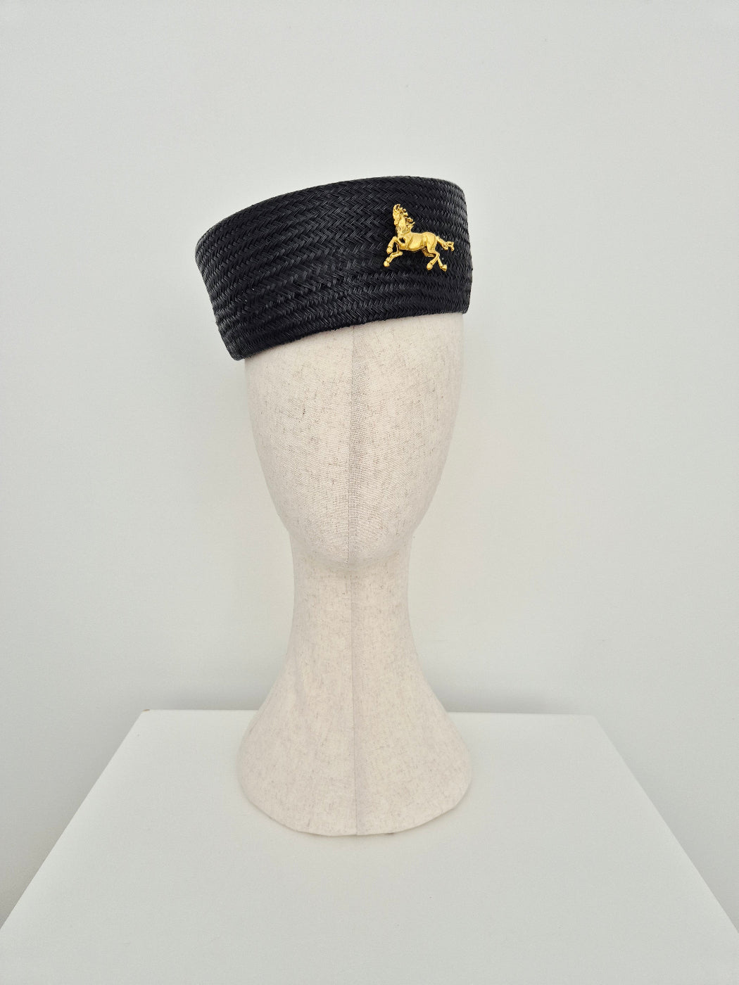 A black woven straw pillbox with a small gold horse emblem on the side, displayed on a cream-colored mannequin head against a light gray background.