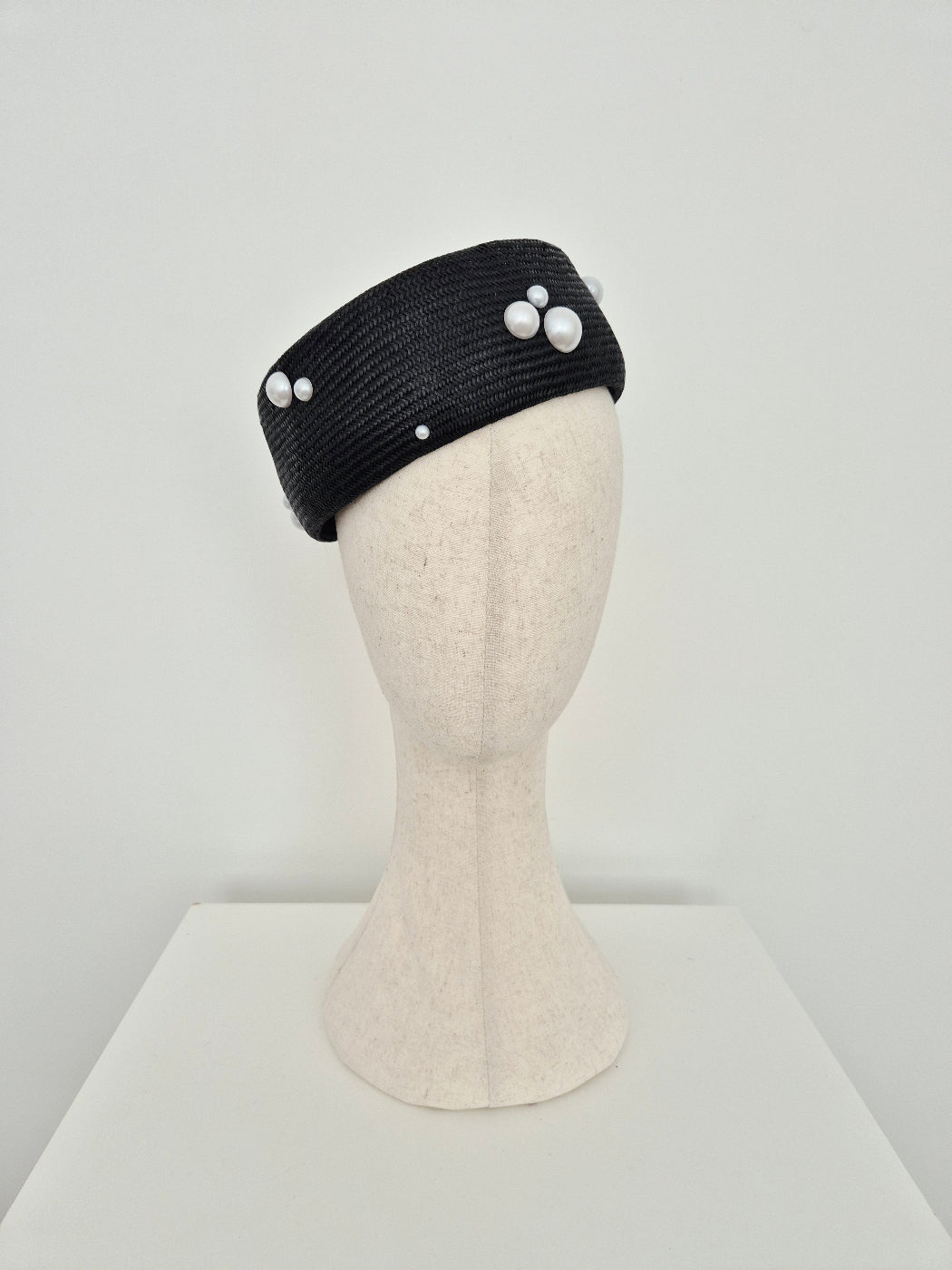 A black straw fascinator headband with white faux-pearl accents, displayed on a cream-colored mannequin head against a neutral light gray background.tilted