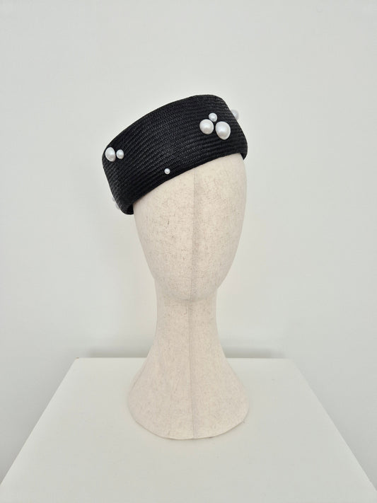 A black straw fascinator headband with white faux-pearl accents, displayed on a cream-colored mannequin head against a neutral light gray background.tilted