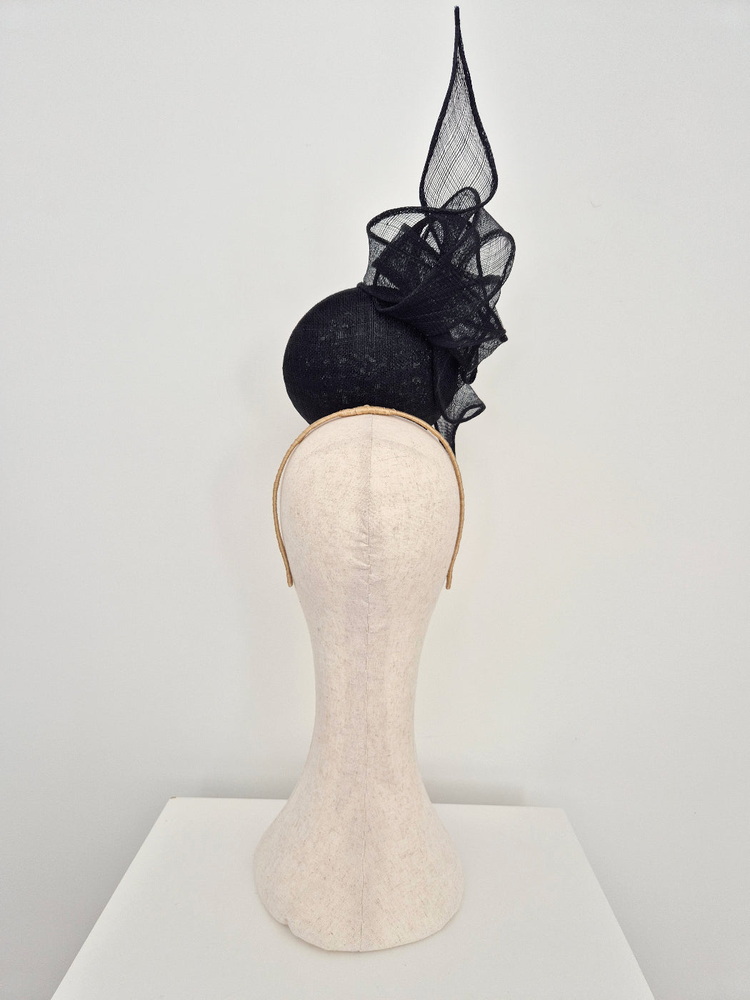 A black straw fascinator percher with a tall pointed knotted tail displayed on a cream-colored mannequin head and neck stand against a plain white background. Back view