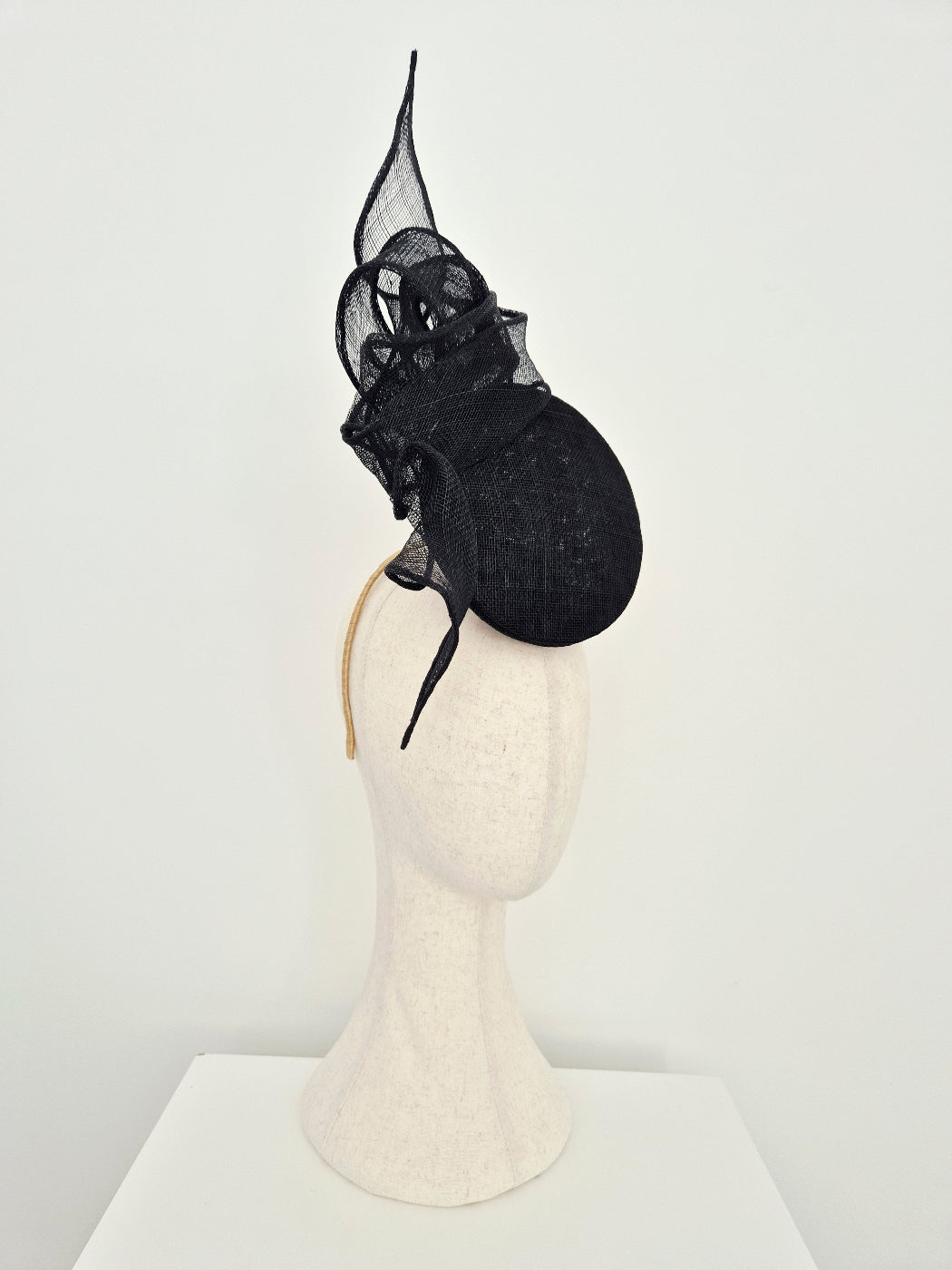 A black straw percher fascinator  with a tall pointed knotted detail and a round structured base, displayed on a cream-colored mannequin head against a plain white background. side view